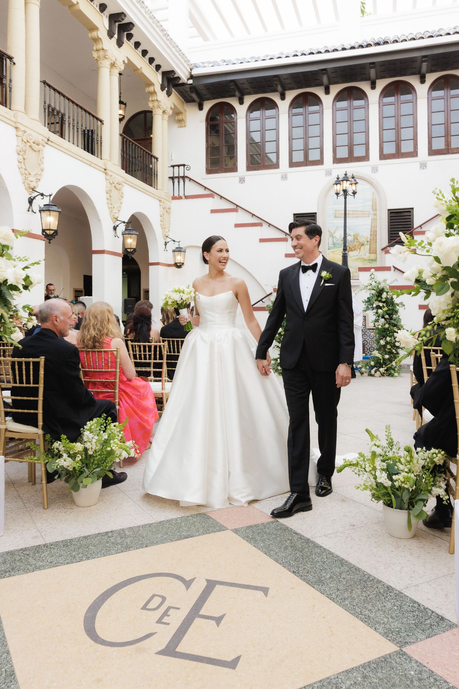 Wedding party gathers in an elegant white columned courtyard with decorative arches and greenery.