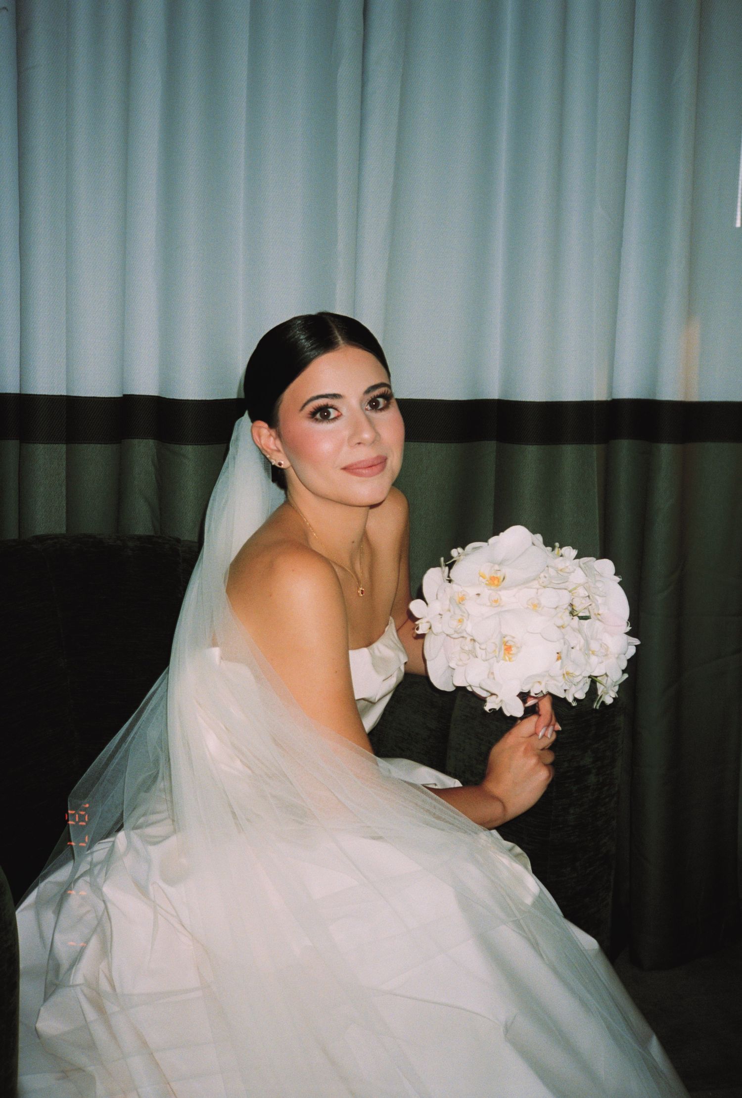 Bride in white strapless gown and veil holds white peony bouquet, seated elegantly before blue curtains.