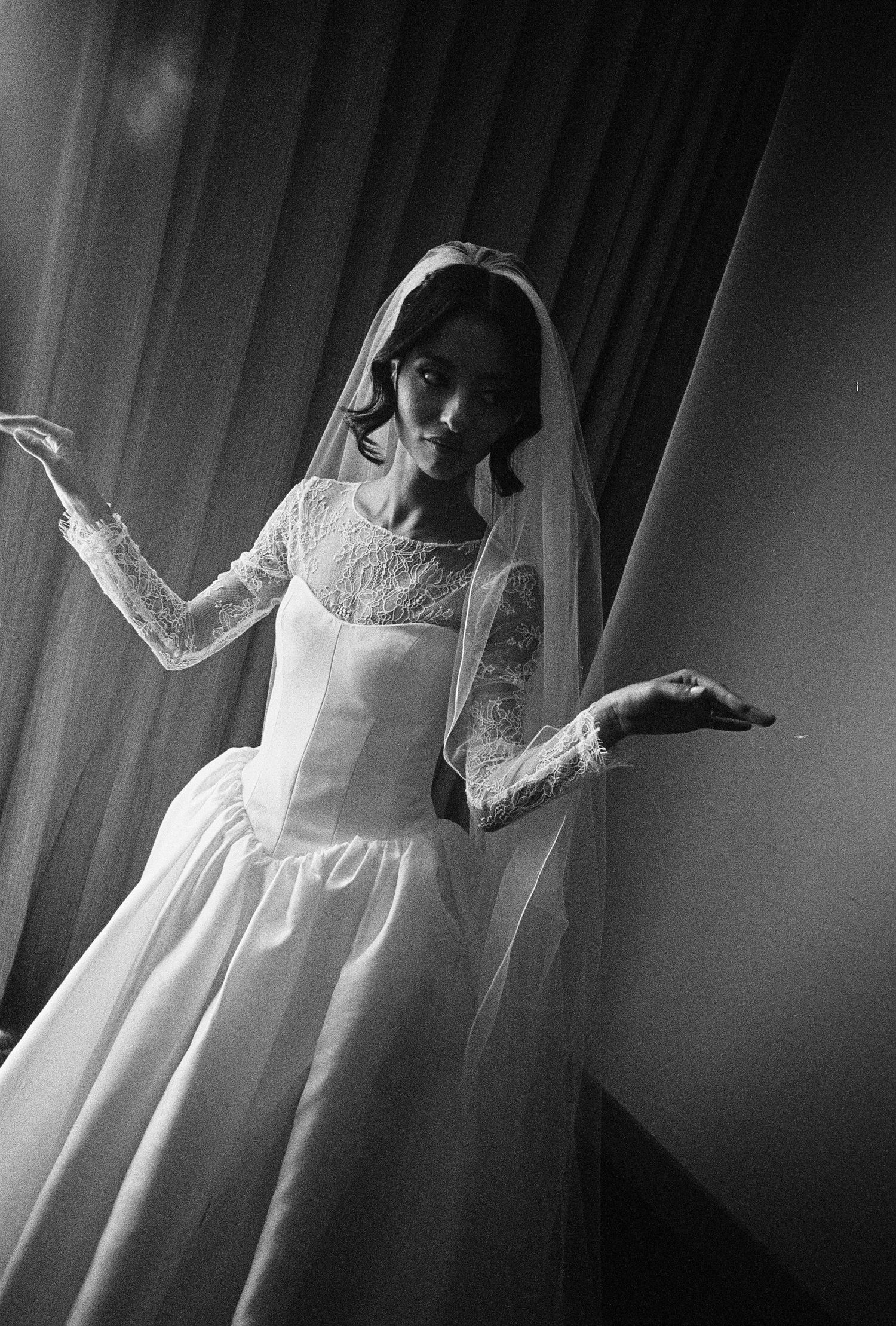 Black and white photo of a young woman in a white lace wedding gown and veil, posing gracefully by a curtain.