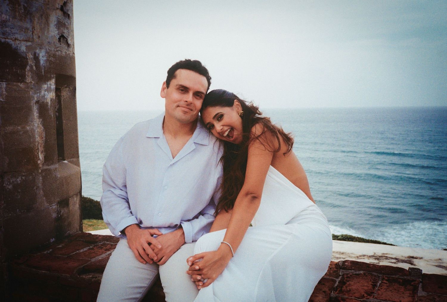 Couple posing together by ocean overlook, woman in white dress leaning on man in light blue shirt, scenic sea backdrop.