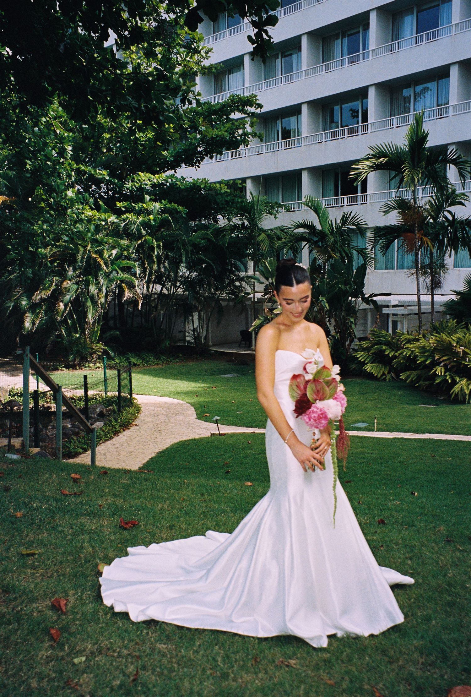 Bride in a strapless white mermaid gown holding pink flowers in a lush hotel garden in color photo.