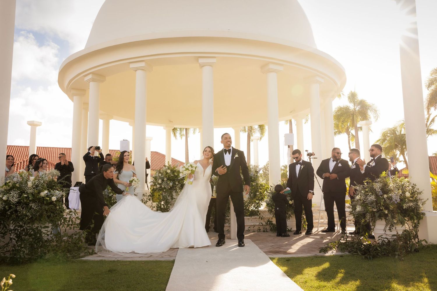 Wedding party gathers under elegant white columned gazebo in lush garden setting at sunset.