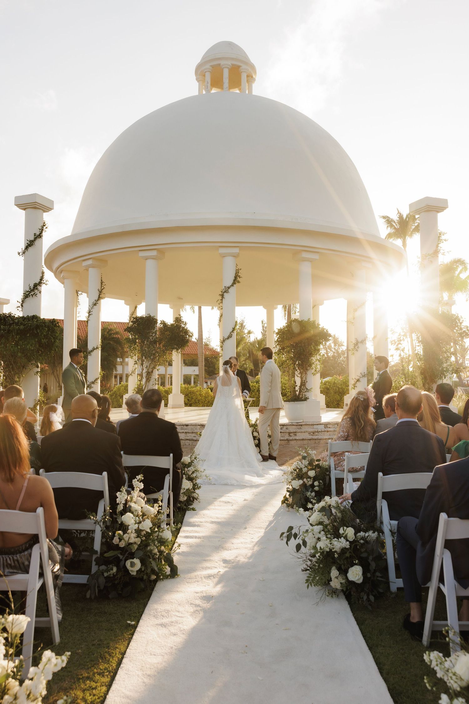 Wedding guests seated in white chairs at outdoor gazebo ceremony during golden hour sunset.