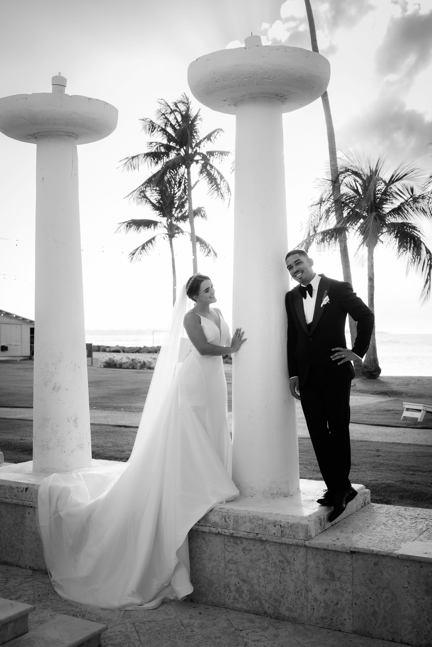 A romantic wedding photo series with a couple posing between elegant white columns against a tropical palm tree backdrop.
