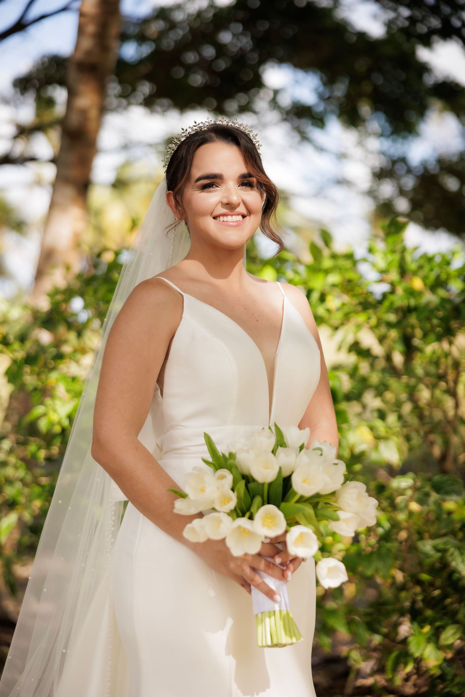 A bride in a white wedding dress holds a bouquet of white flowers against a sunlit natural outdoor backdrop with greenery.