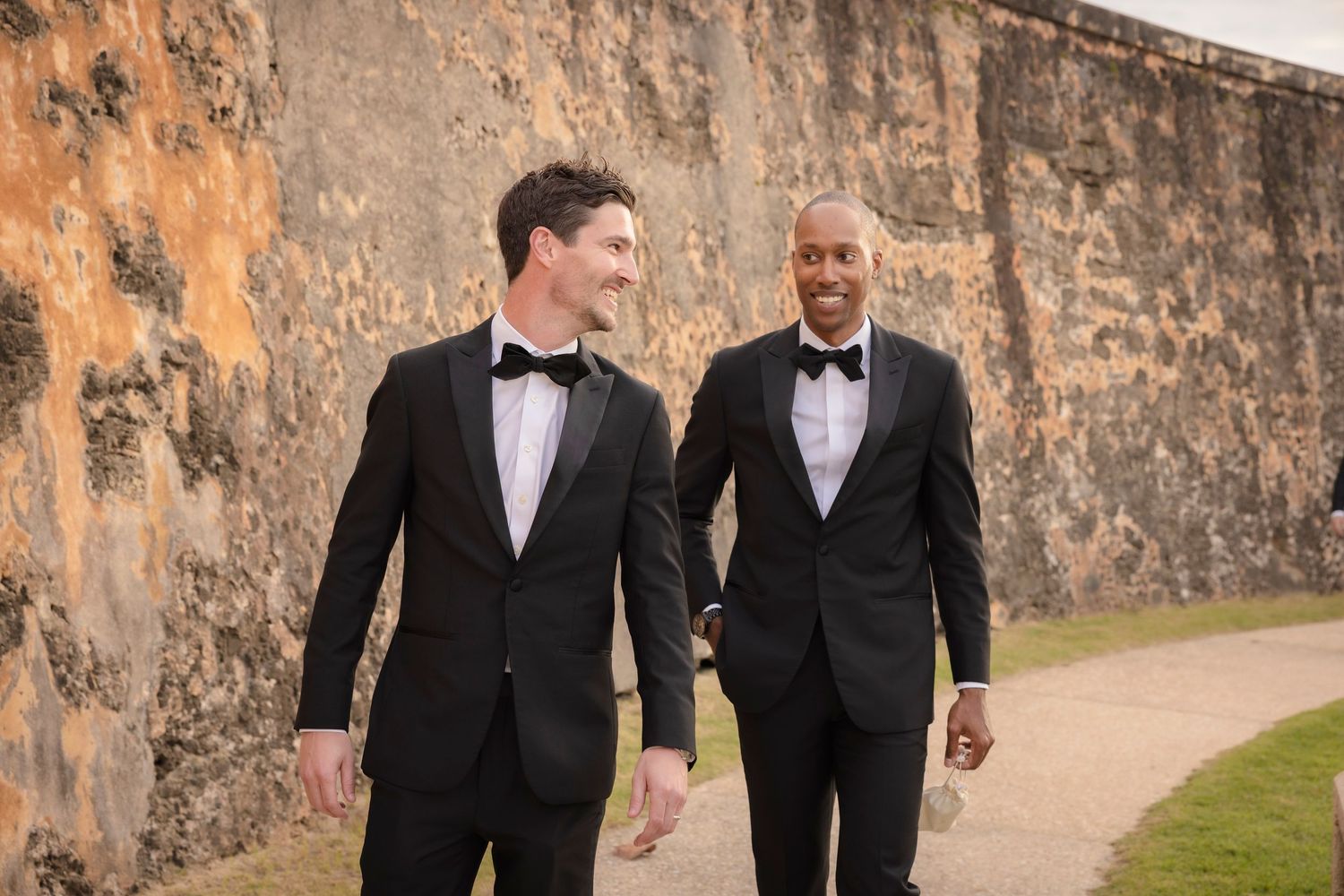 Groomsmen in black tuxedos walk along historic fortress wall during wedding.