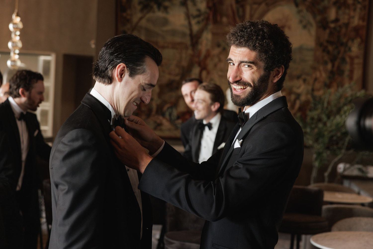Wedding party members in formal black tuxedos share candid moments in a warmly lit venue.