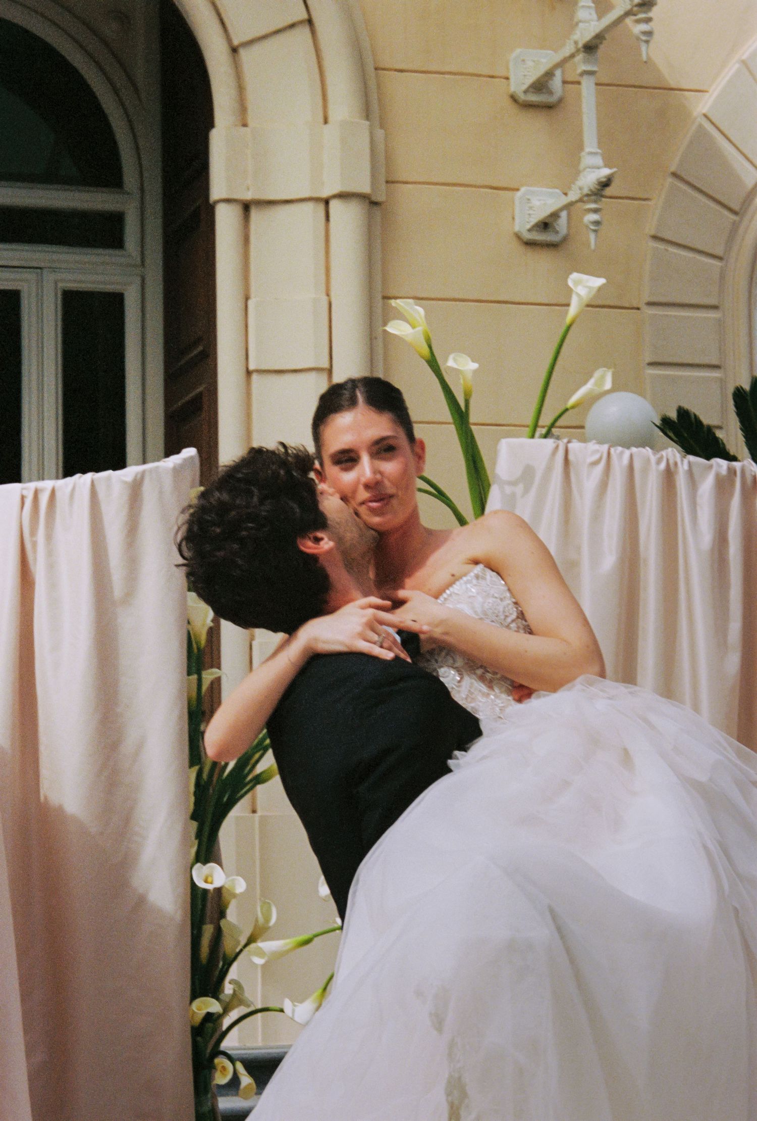 Bride and groom sharing a romantic kiss at their wedding, surrounded by calla lilies and blush pink draping.