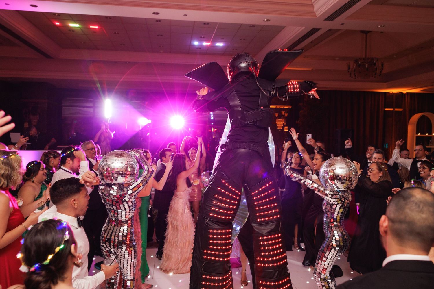 Party guests dance and celebrate on a lively dance floor illuminated with vibrant pink and purple lighting.