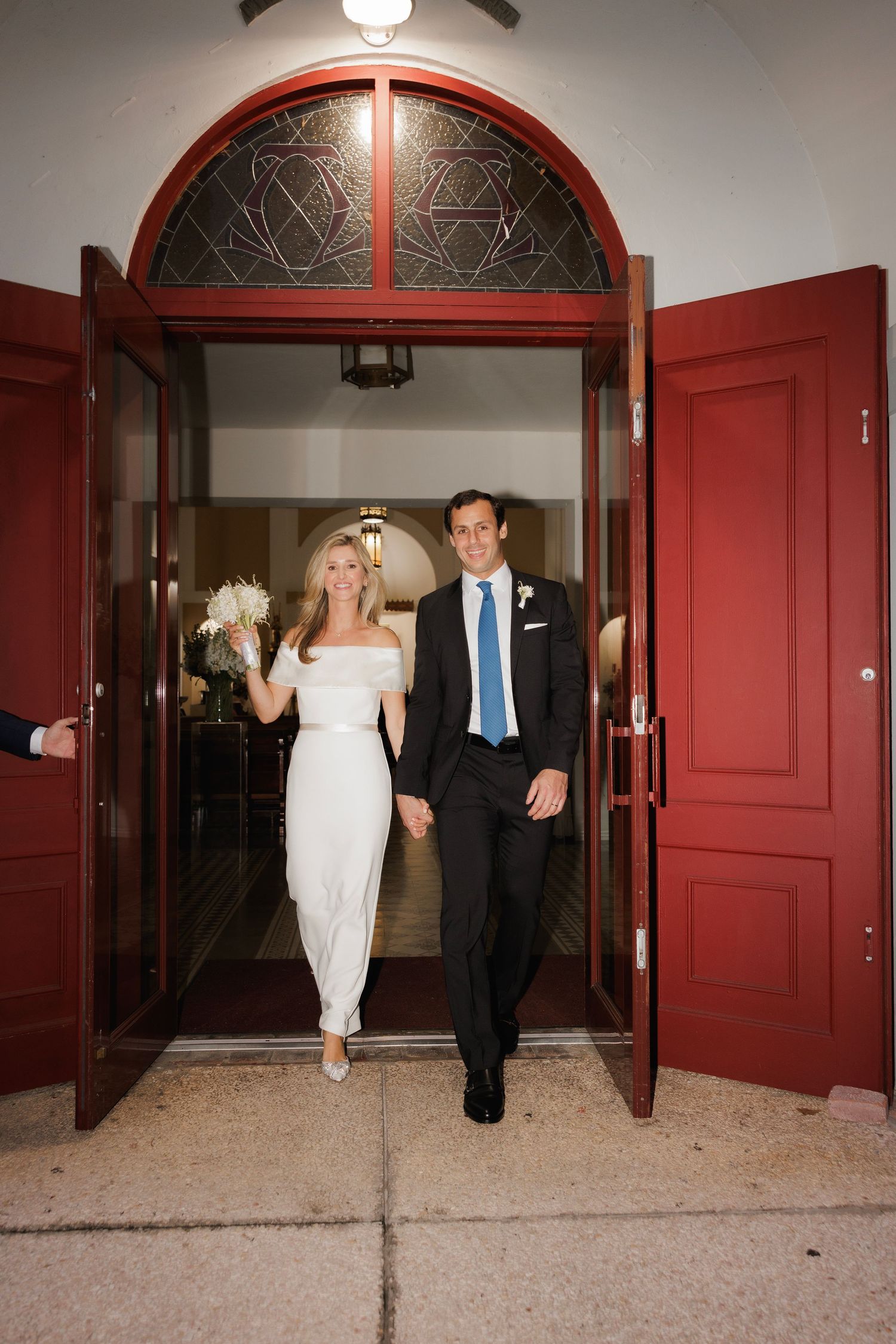 A couple exits through grand red church doors with arched stained glass detail during their wedding celebration.