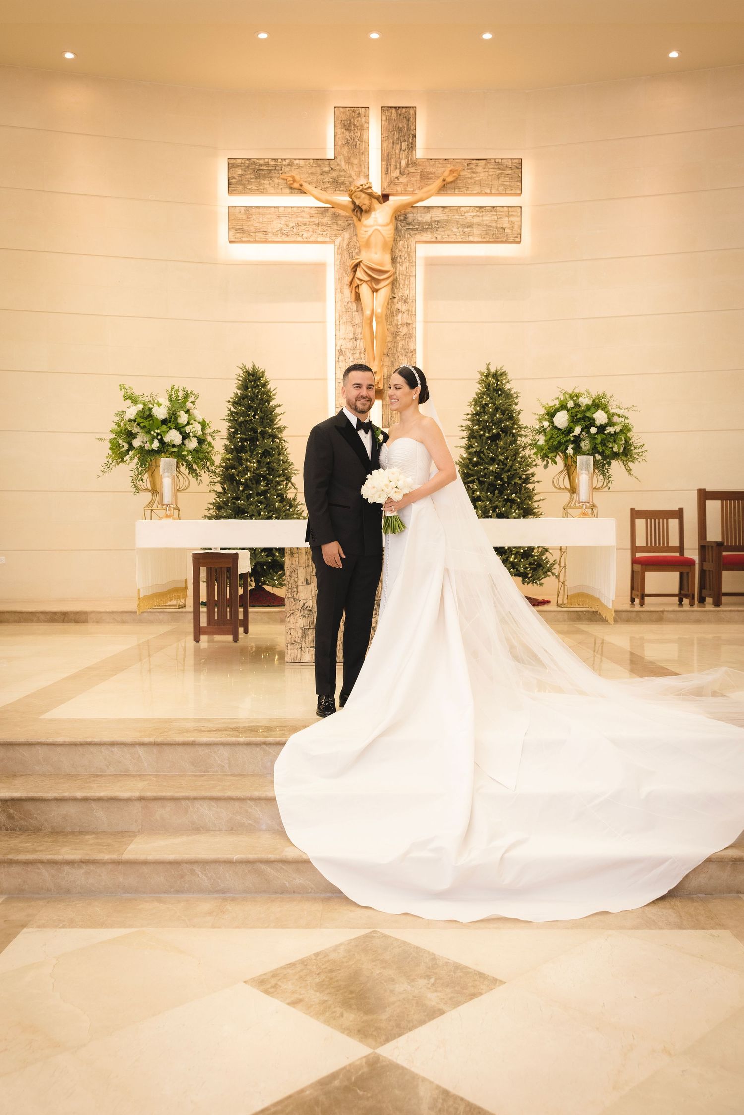 A wedding couple stands in front of a large illuminated crucifix in a church with white floral arrangements and greenery.