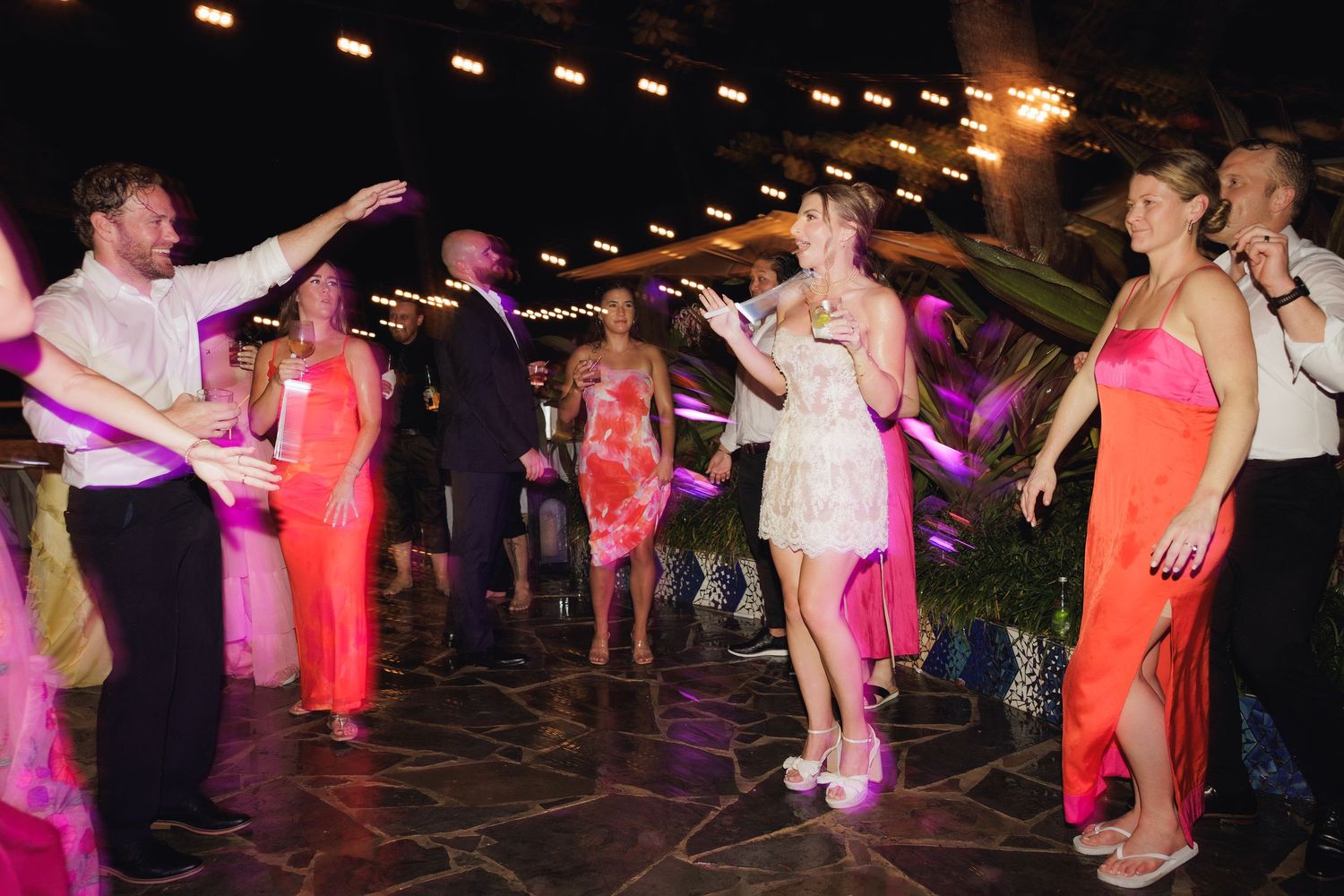 Party guests dance and celebrate on a dark dance floor illuminated by pink and orange lighting.