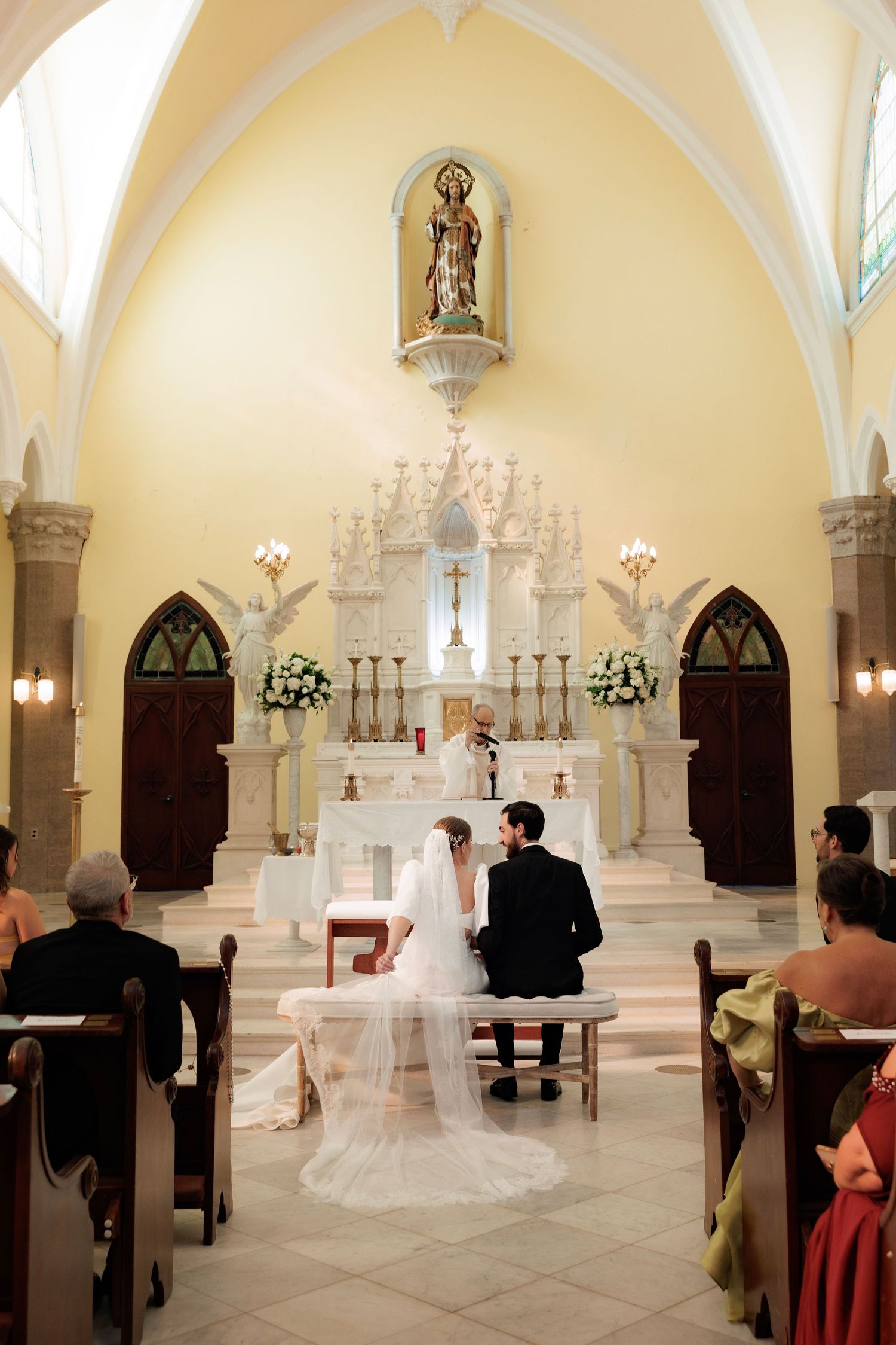 A wedding ceremony takes place in a beautiful white church with vaulted ceilings and gothic arches.