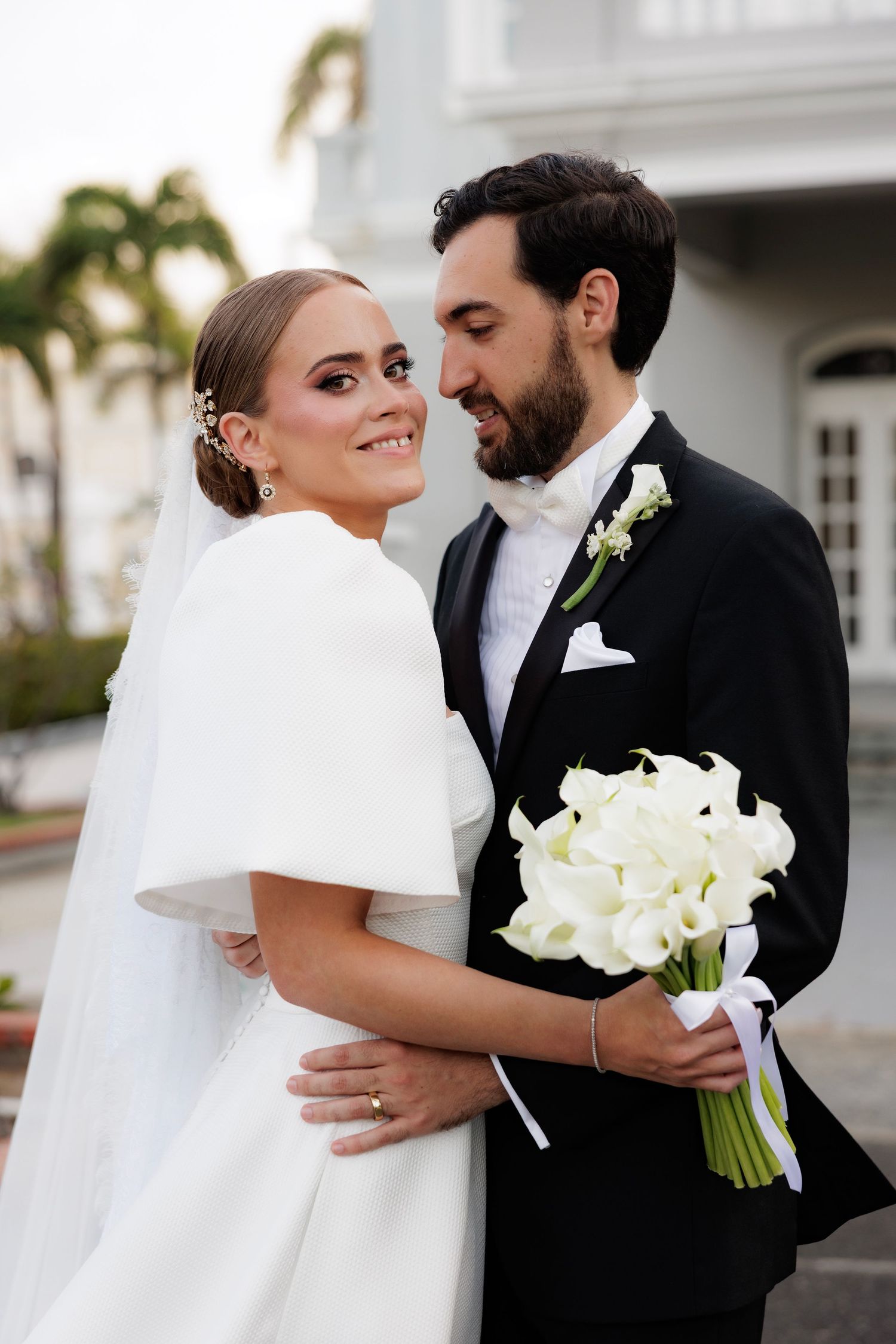 A joyful bride in a white dress with fur stole holds a calla lily bouquet while embracing her groom in a black tuxedo.