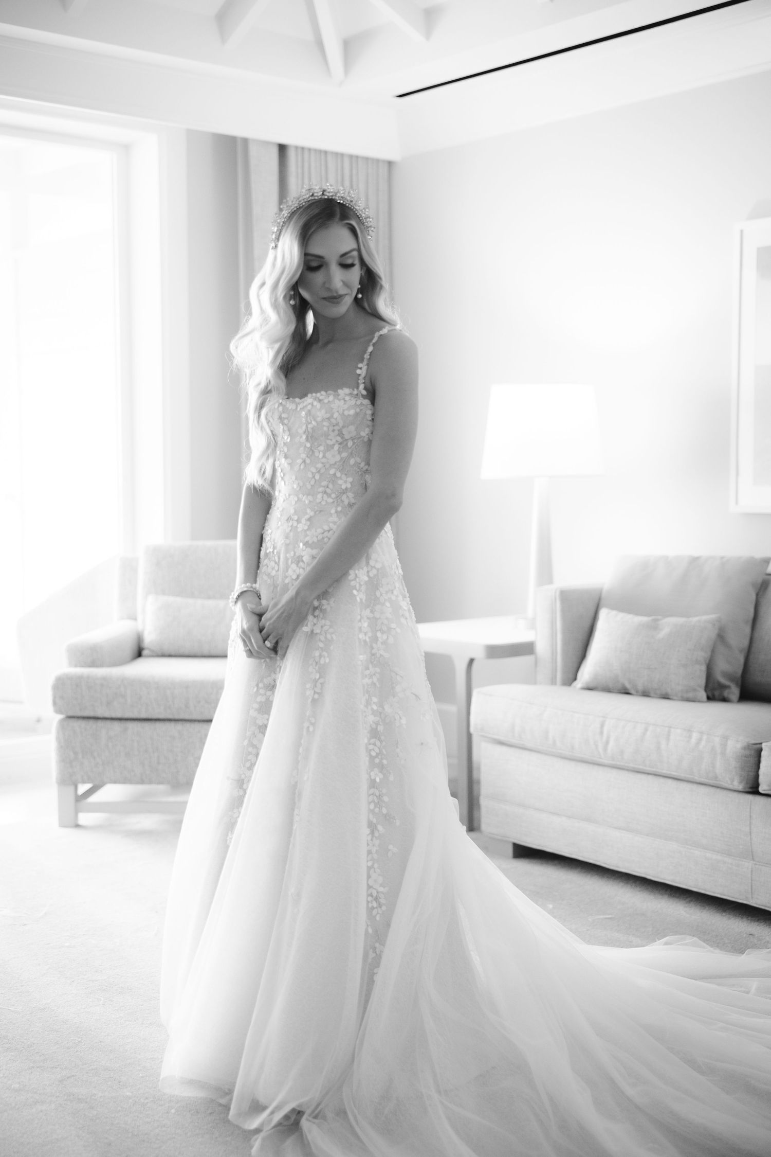 Black and white photo of a bride in a lace wedding dress with long train standing in a bright modern room.