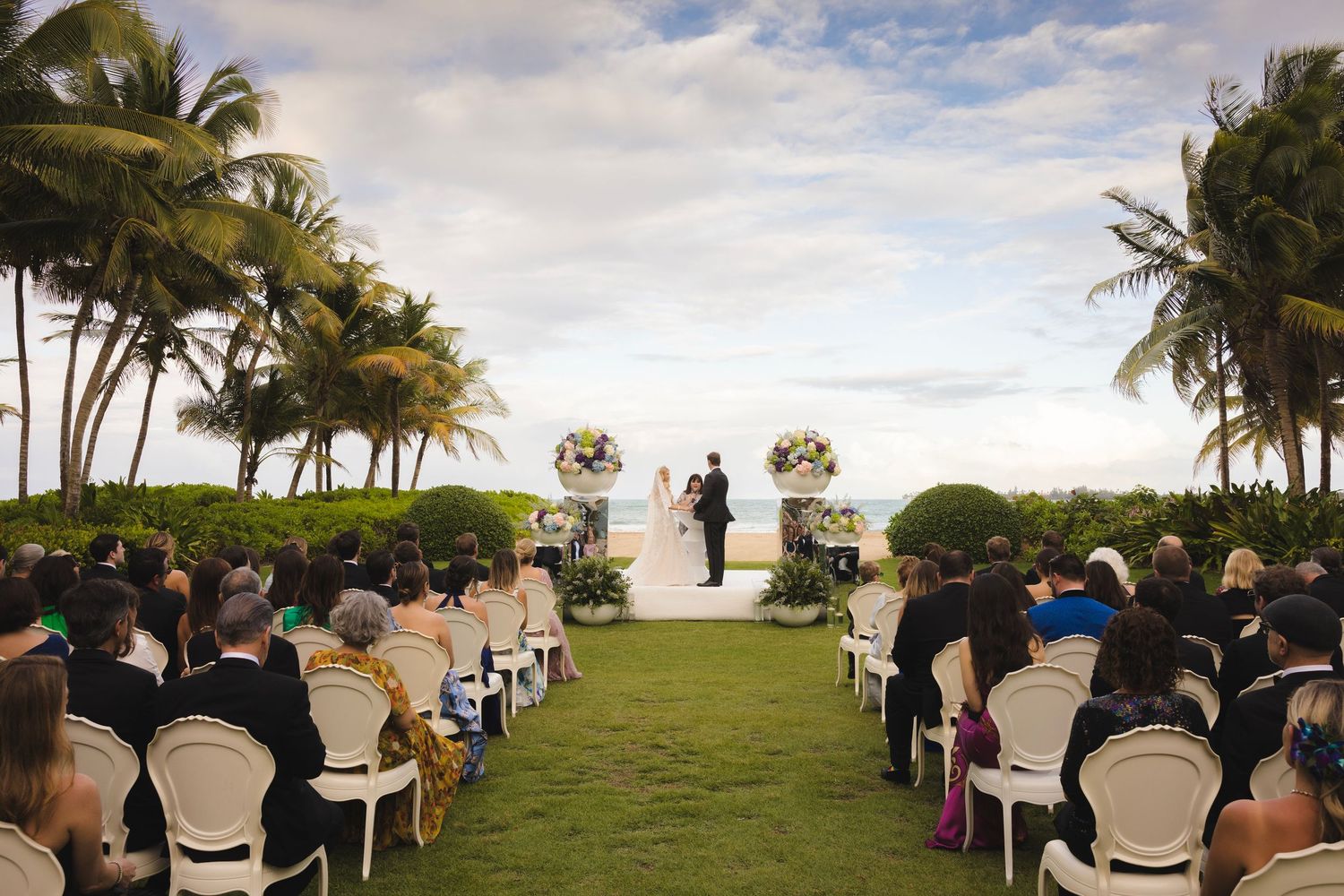 Wedding ceremony setup on a tropical beach with rows of white chairs and palm trees swaying in the breeze.