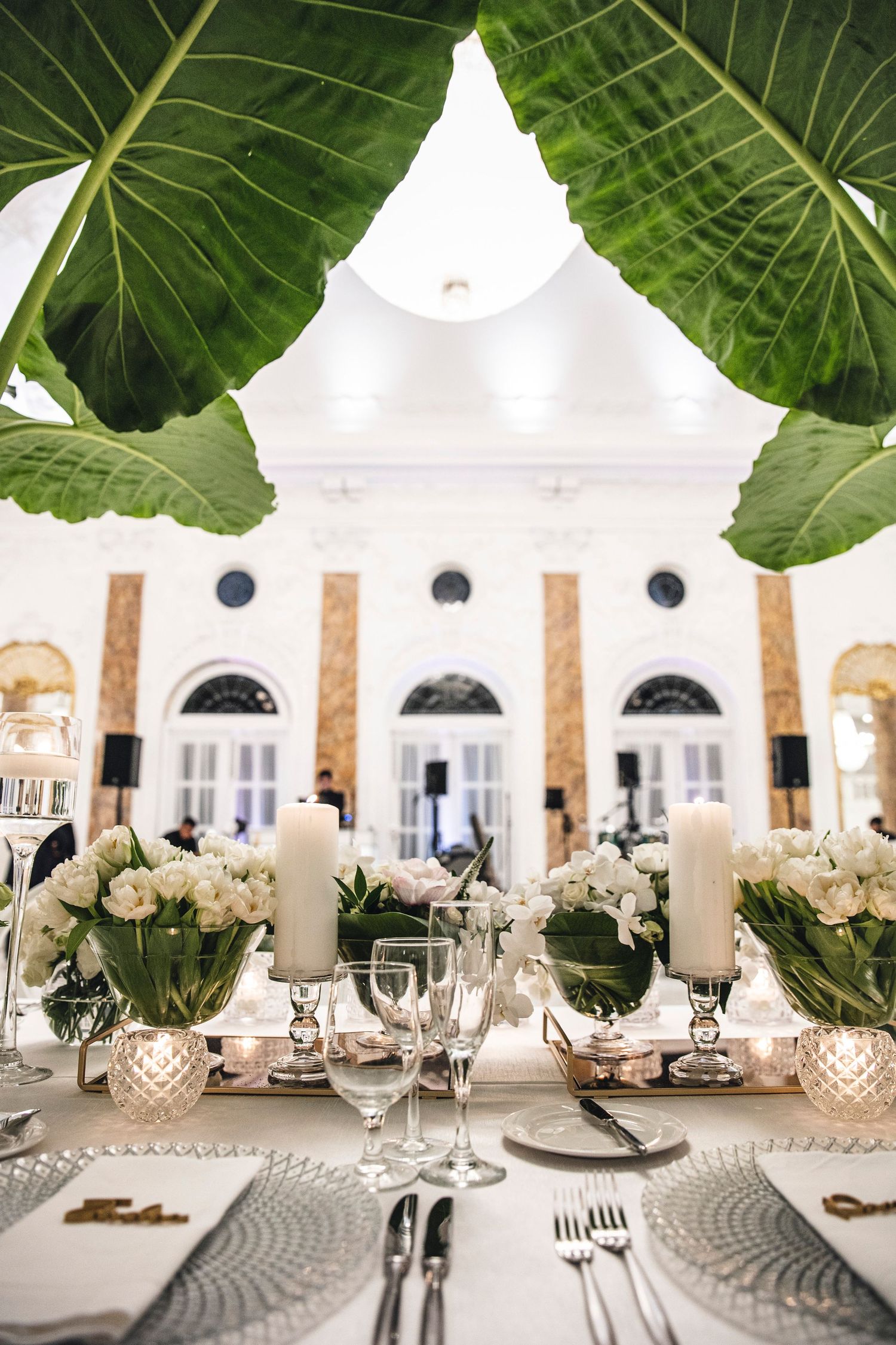 Elegant tropical-themed wedding reception table with large palm leaves and white floral centerpieces in an ornate ballroom.