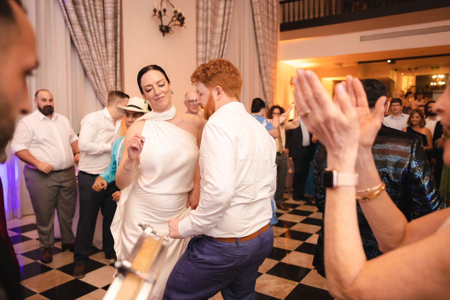 Wedding guests dance enthusiastically on a black and white checkered dance floor at an evening reception.