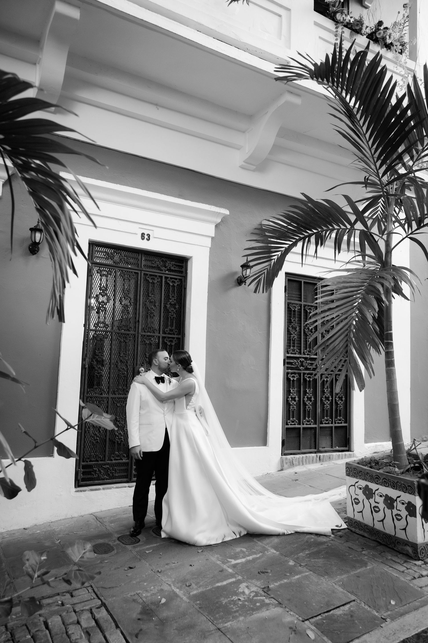 A romantic wedding photo in black and white showing a couple embracing in front of a colonial style building with palm trees.