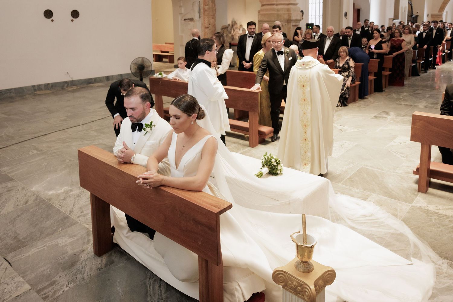 A couple sits in a wooden church pew during a traditional Catholic wedding ceremony with clergy and guests visible.