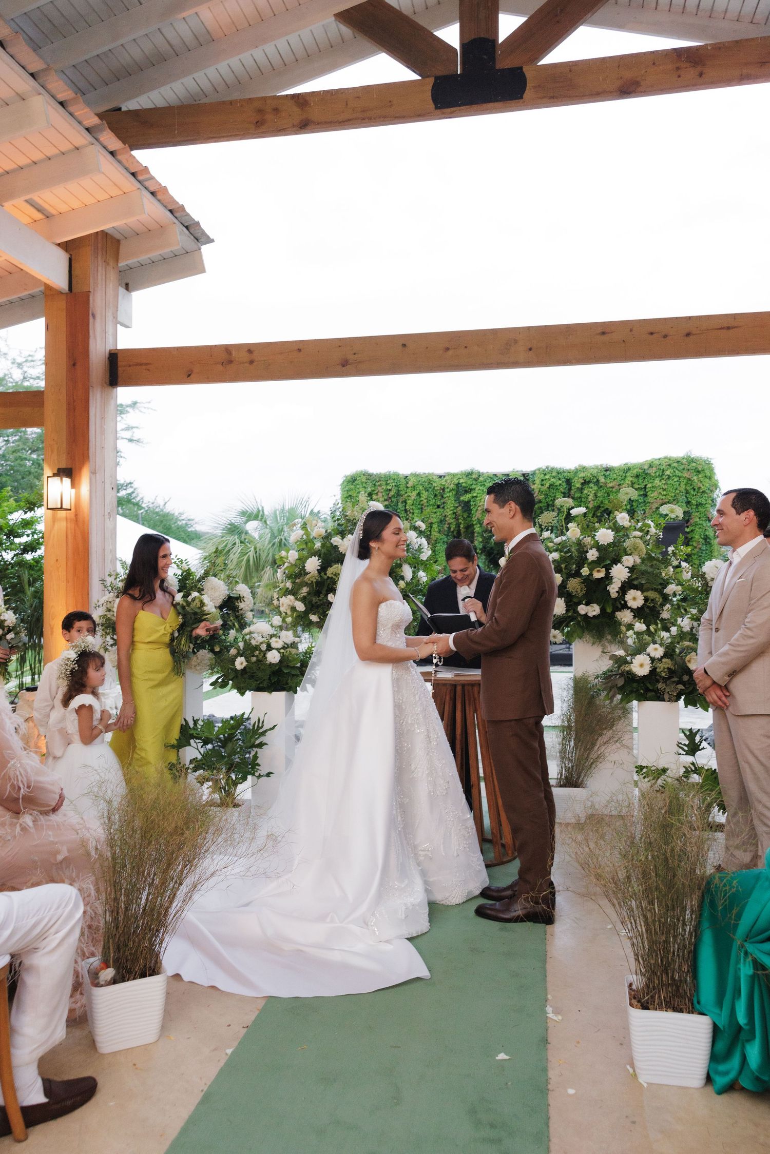 A wedding ceremony takes place under a wooden pergola with white floral arrangements and guests in colorful attire.