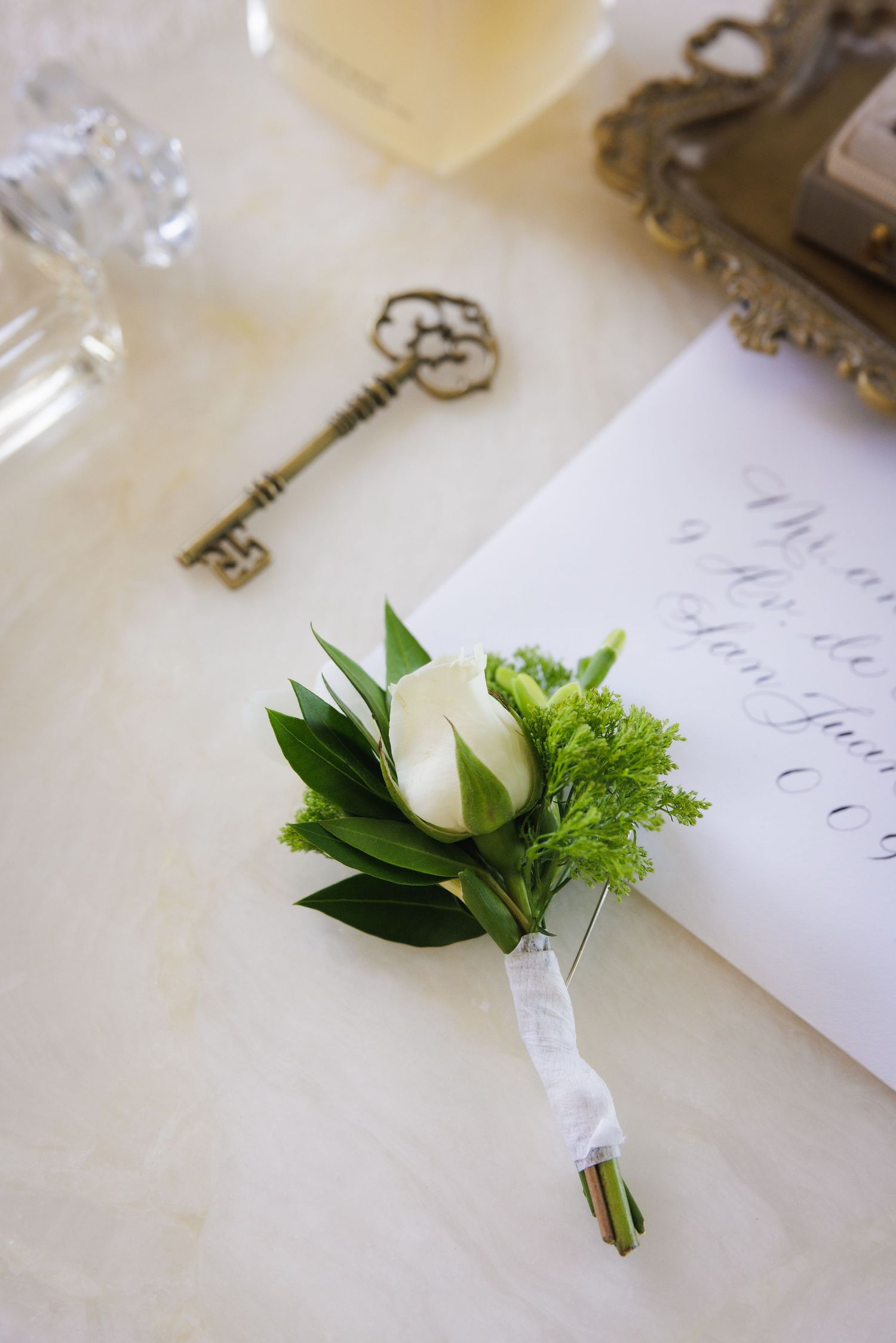 Elegant white rose boutonniere with greenery next to decorative antique key and handwritten note.