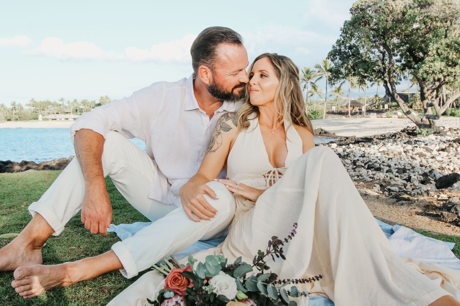 Couple sitting on grass at Kaikaua Point Beach Park Kona Hawaii with groom kissing bride’s cheek at sunset overlooking ocean