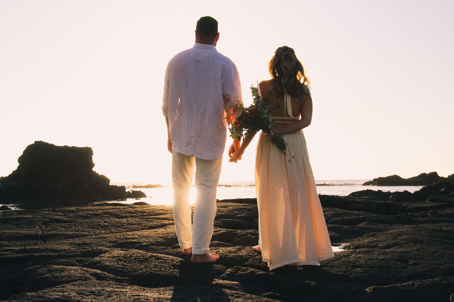 Couple standing on rocks at sunset at Kaikaua Point Beach Park Kona Hawaii with bride facing camera and groom looking toward ocean