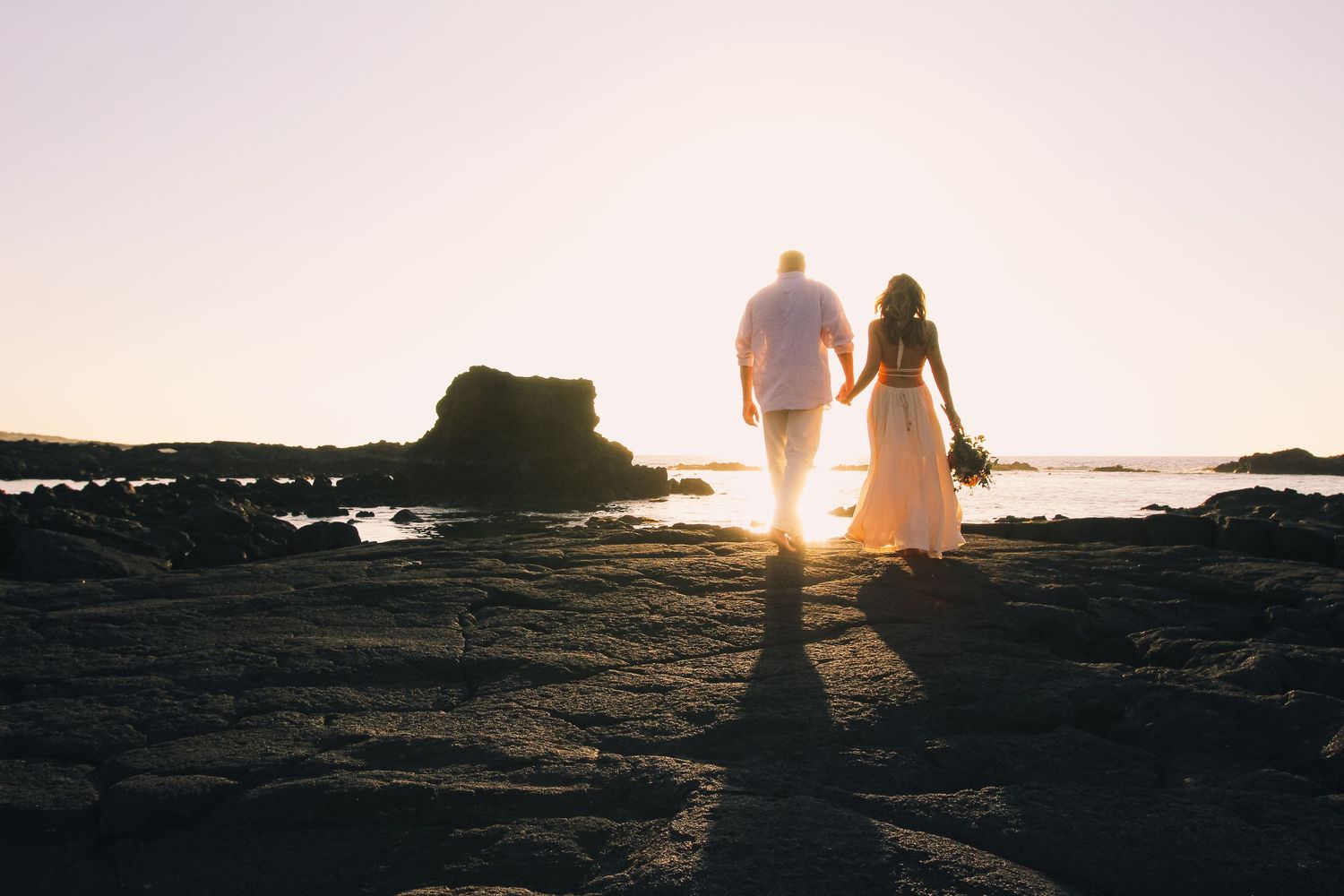 Couple holding hands on rocks at sunset at Kaikaua Point Beach Park Kona Hawaii with sun setting over ocean