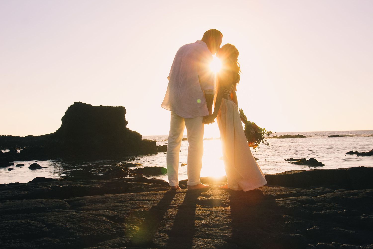 Couple kissing on rocks at sunset at Kaikaua Point Beach Park Kona Hawaii with sun backlighting ocean scene
