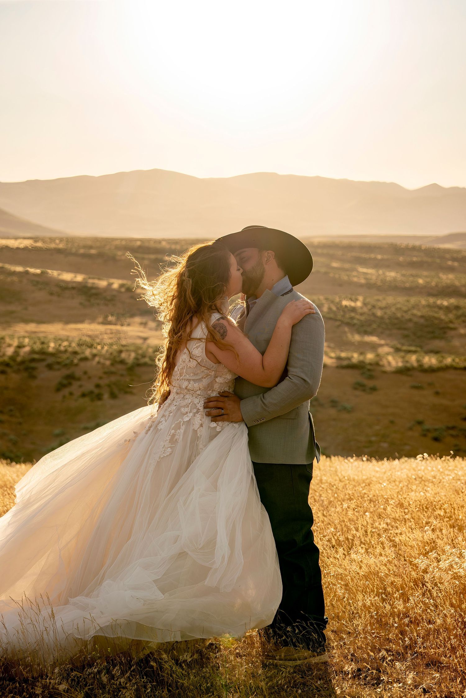 Bride and groom share a passionate kiss on a golden hillside at sunset with mountain views.