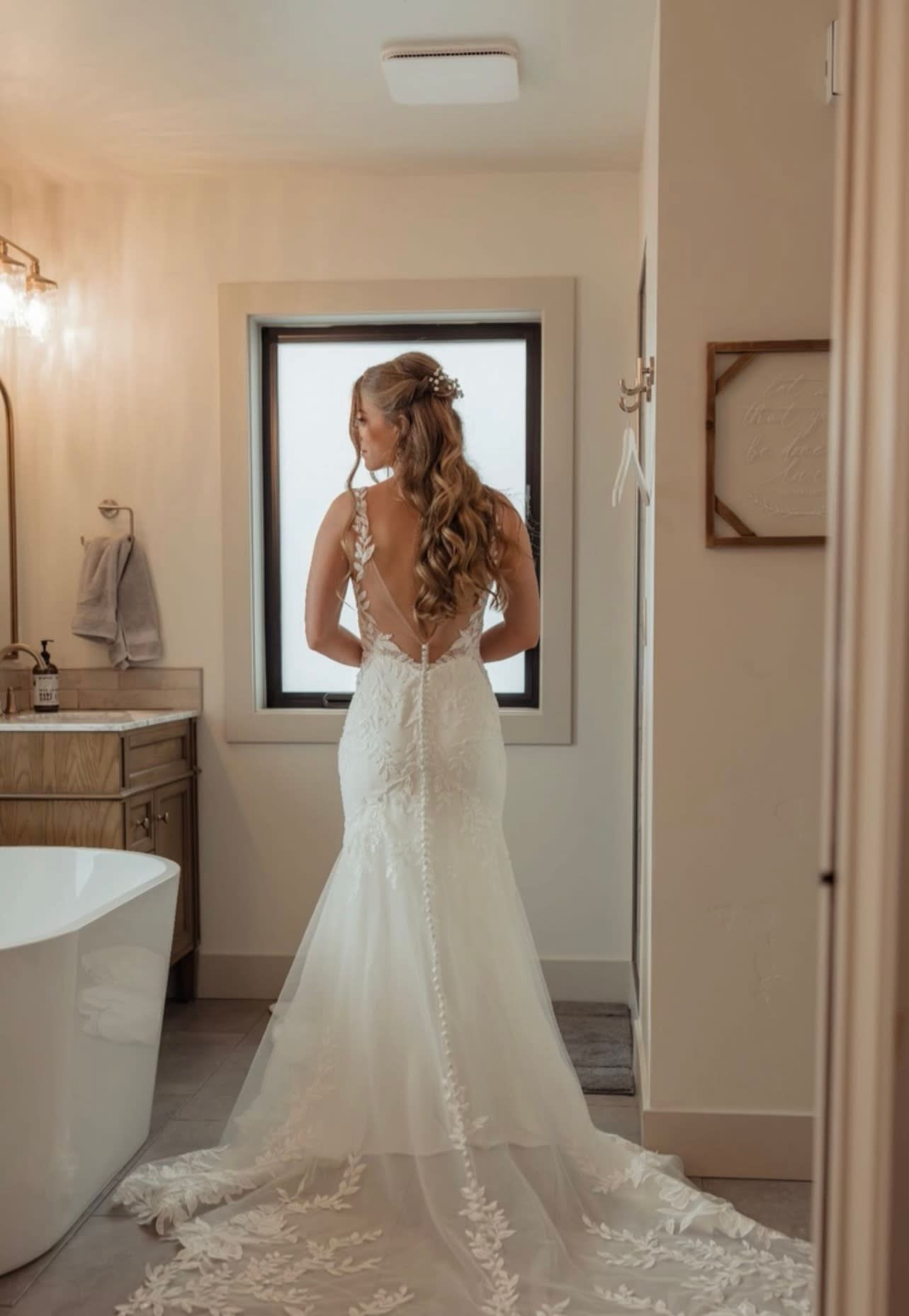 Bride in backless lace wedding gown with train standing by bathroom window.