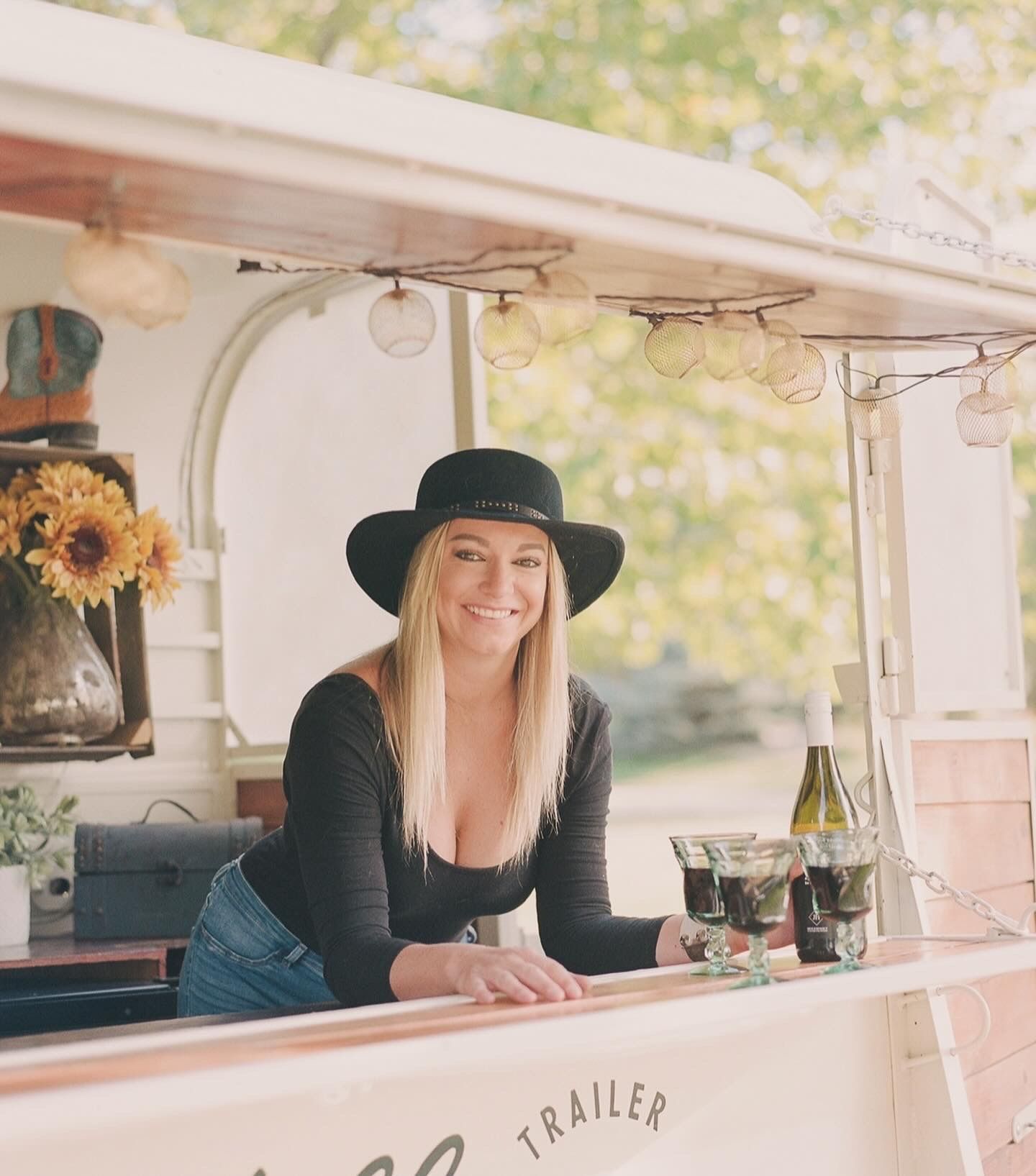 Smiling blonde woman in black hat serving drinks from a vintage mobile bar trailer with sunflowers.