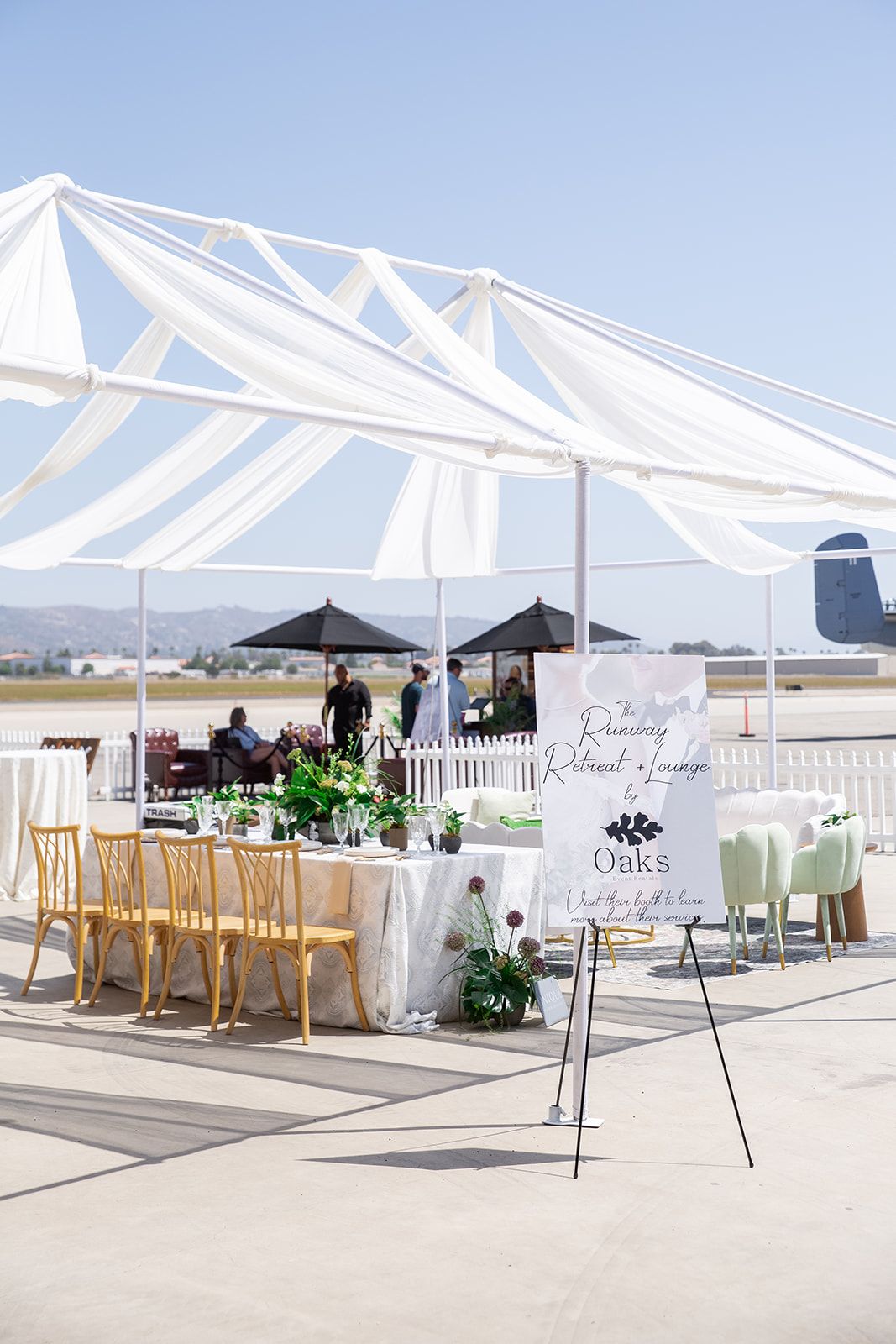 Elegant outdoor wedding expo setup with white draped canopy, gold chairs, and floral displays on an airport tarmac.