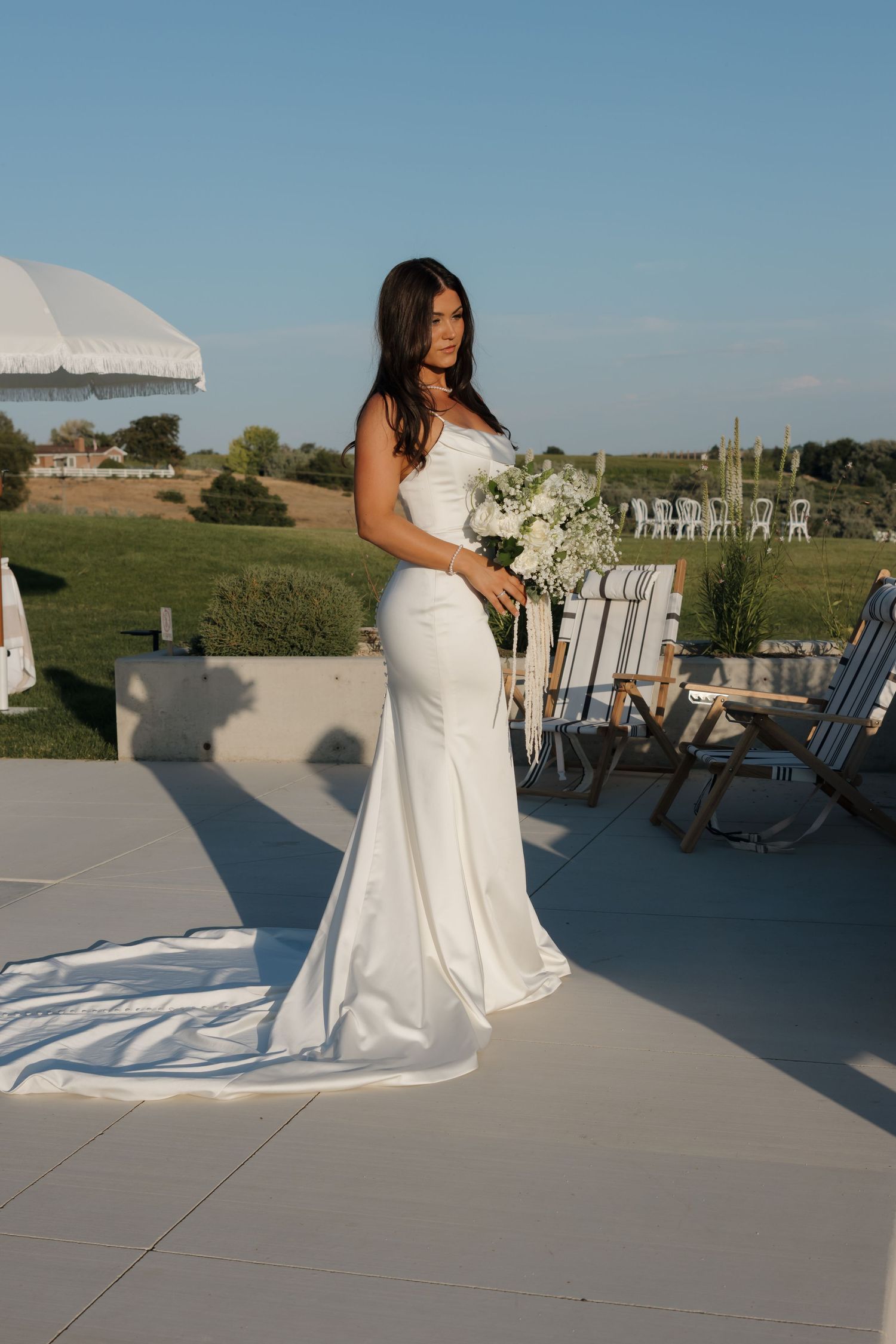 Bride in fitted white satin gown holding white floral bouquet standing on outdoor wedding venue patio.