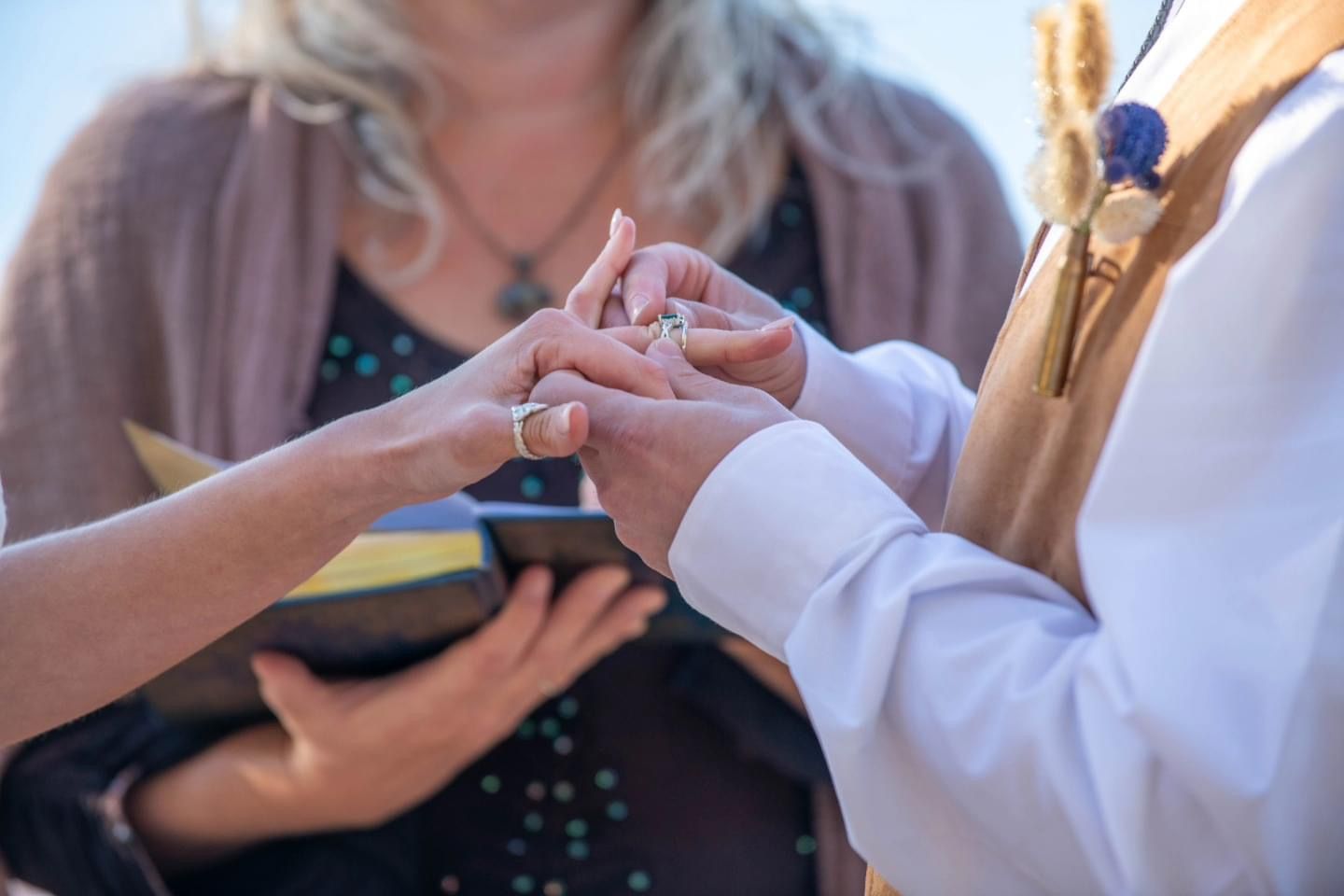 Close-up of wedding ring exchange ceremony with officiant holding book during intimate outdoor wedding ritual.