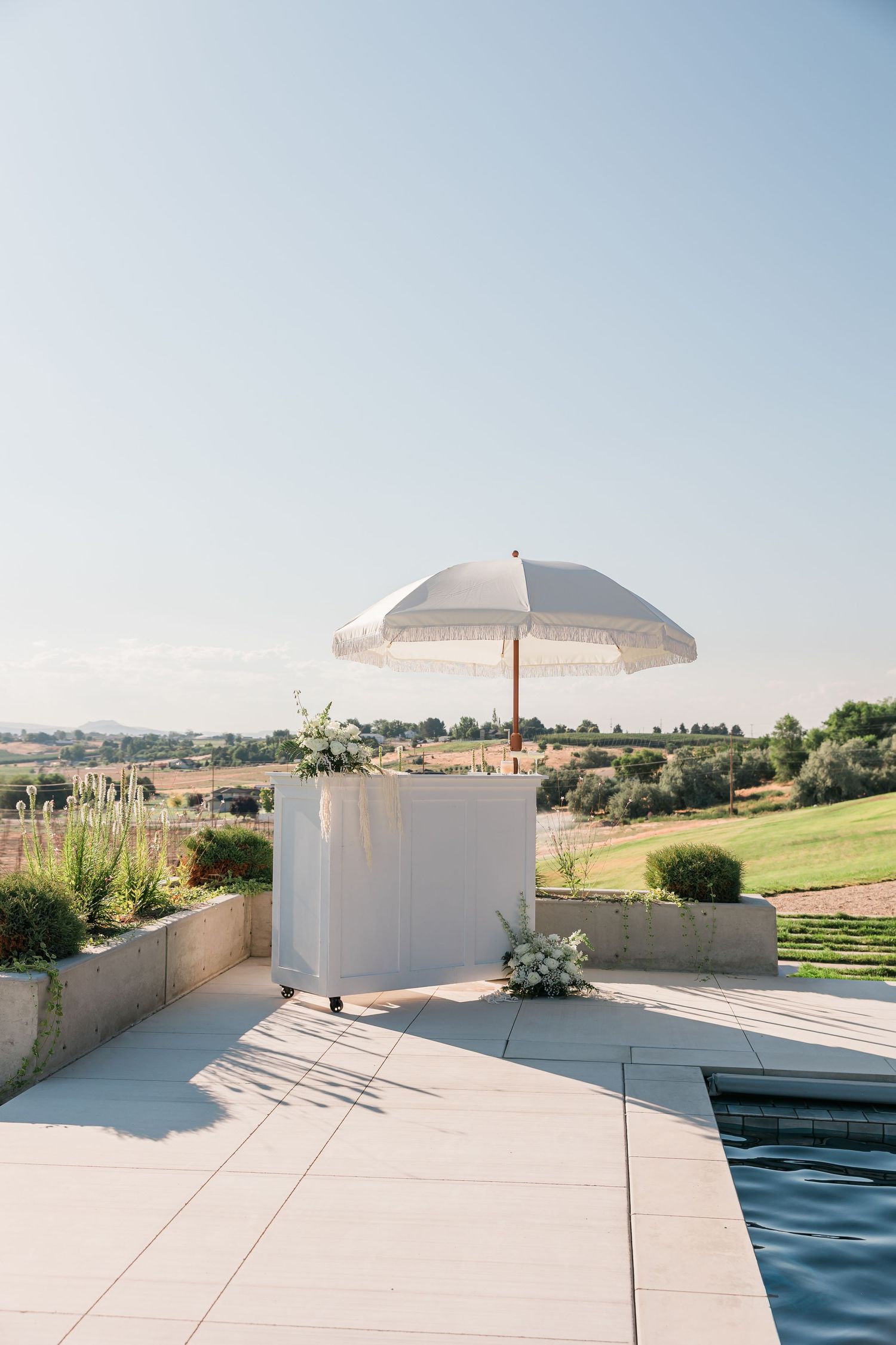 Elegant outdoor wedding bar setup with white umbrella and floral arrangements beside a pool with vineyard views.
