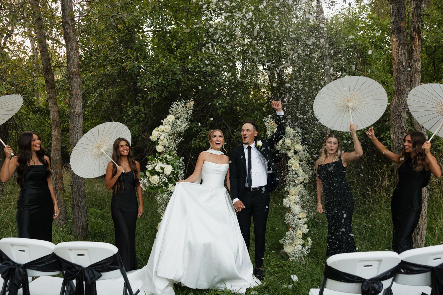 Joyful newlyweds celebrate with bridesmaids holding white parasols amid confetti shower in wooded outdoor venue.