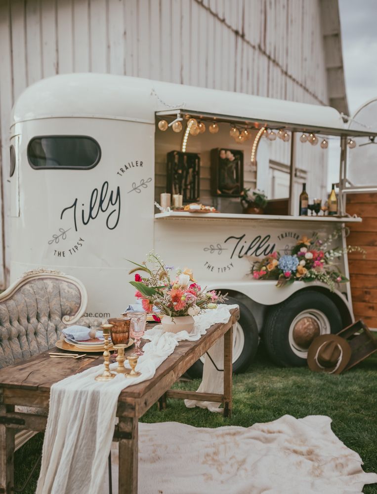 Vintage white Tilly horse trailer bar setup with rustic wooden table and colorful floral centerpiece.