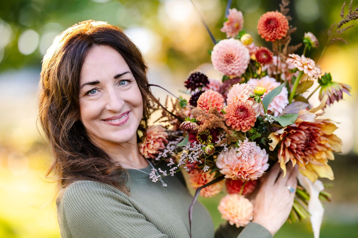 Woman smiling while holding a lush autumn floral arrangement of dahlias outdoors.
