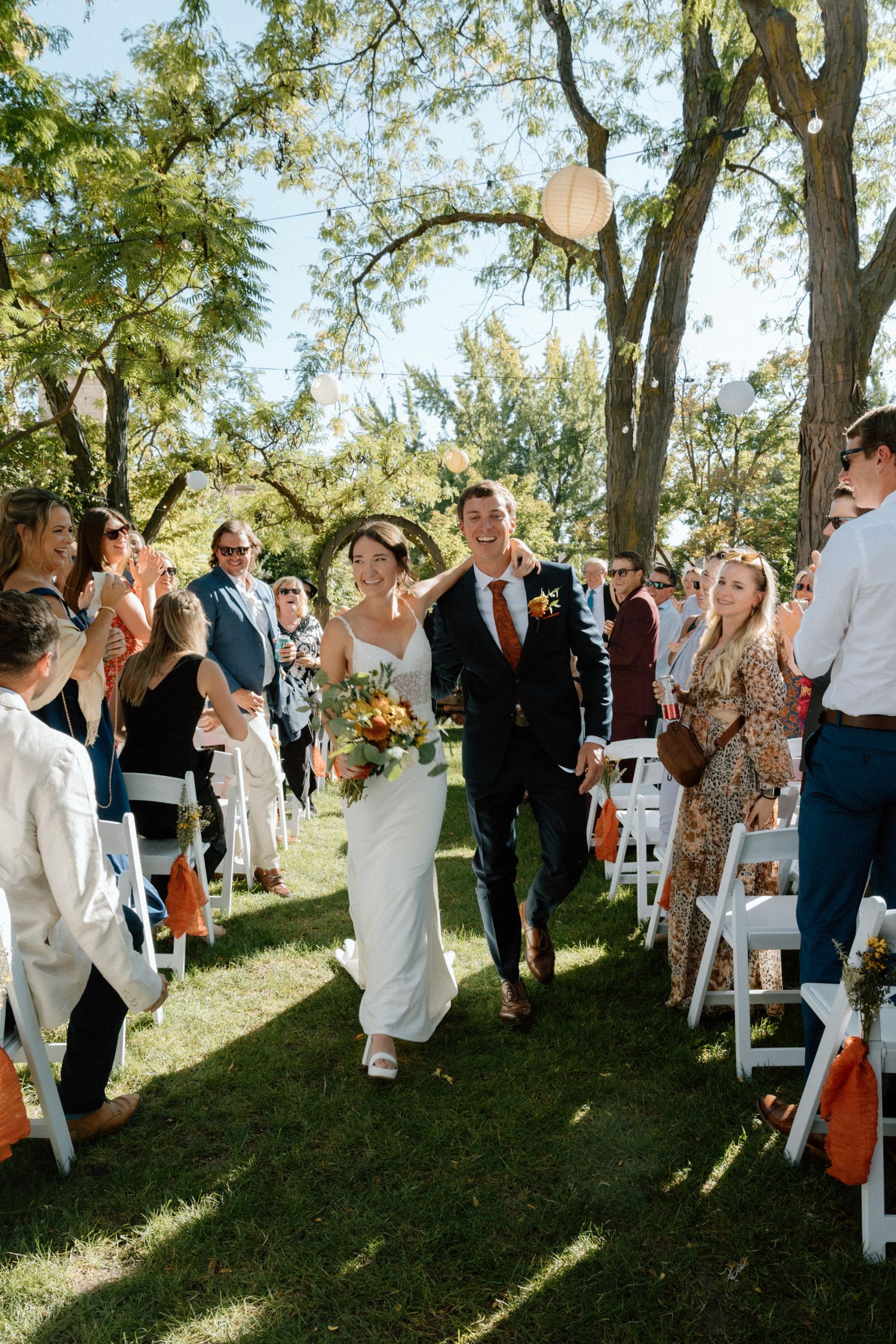 Bride and groom walk down outdoor wedding aisle surrounded by cheering guests under green trees.