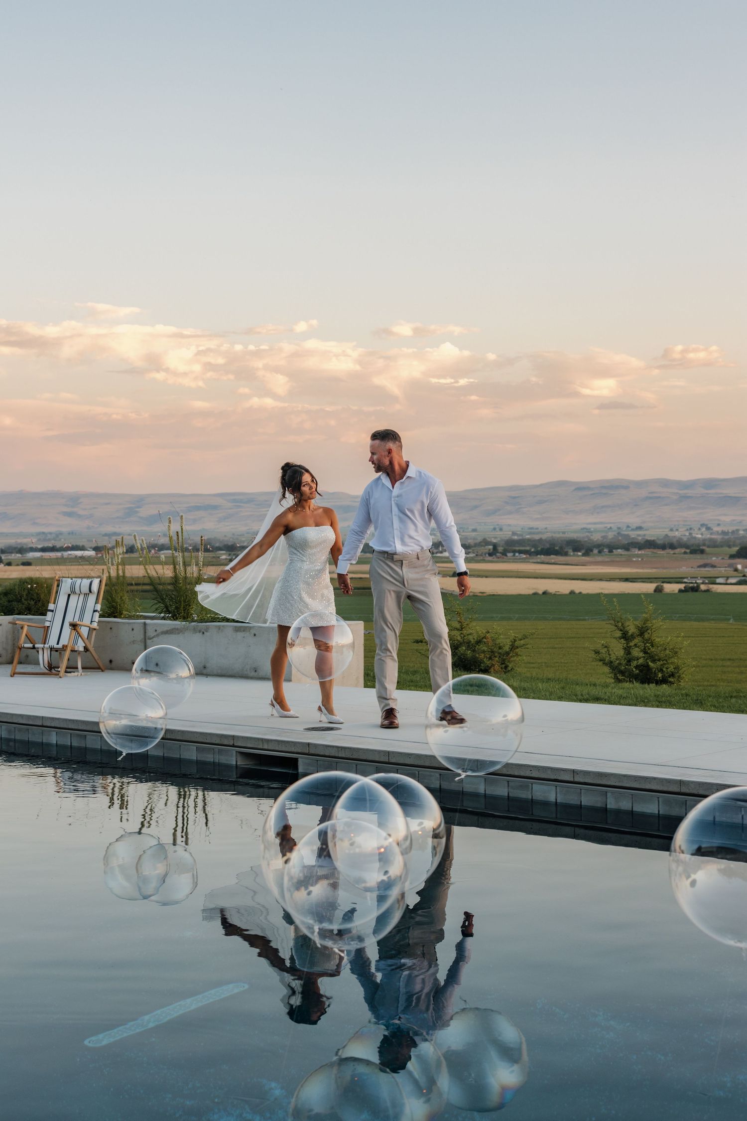 Couple in white holding hands by infinity pool with scenic valley sunset view.