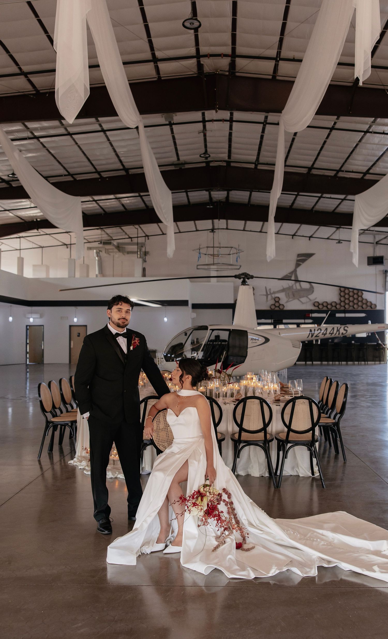 Bride and groom pose elegantly in a decorated airplane hangar wedding venue with a helicopter and candlelit reception table.