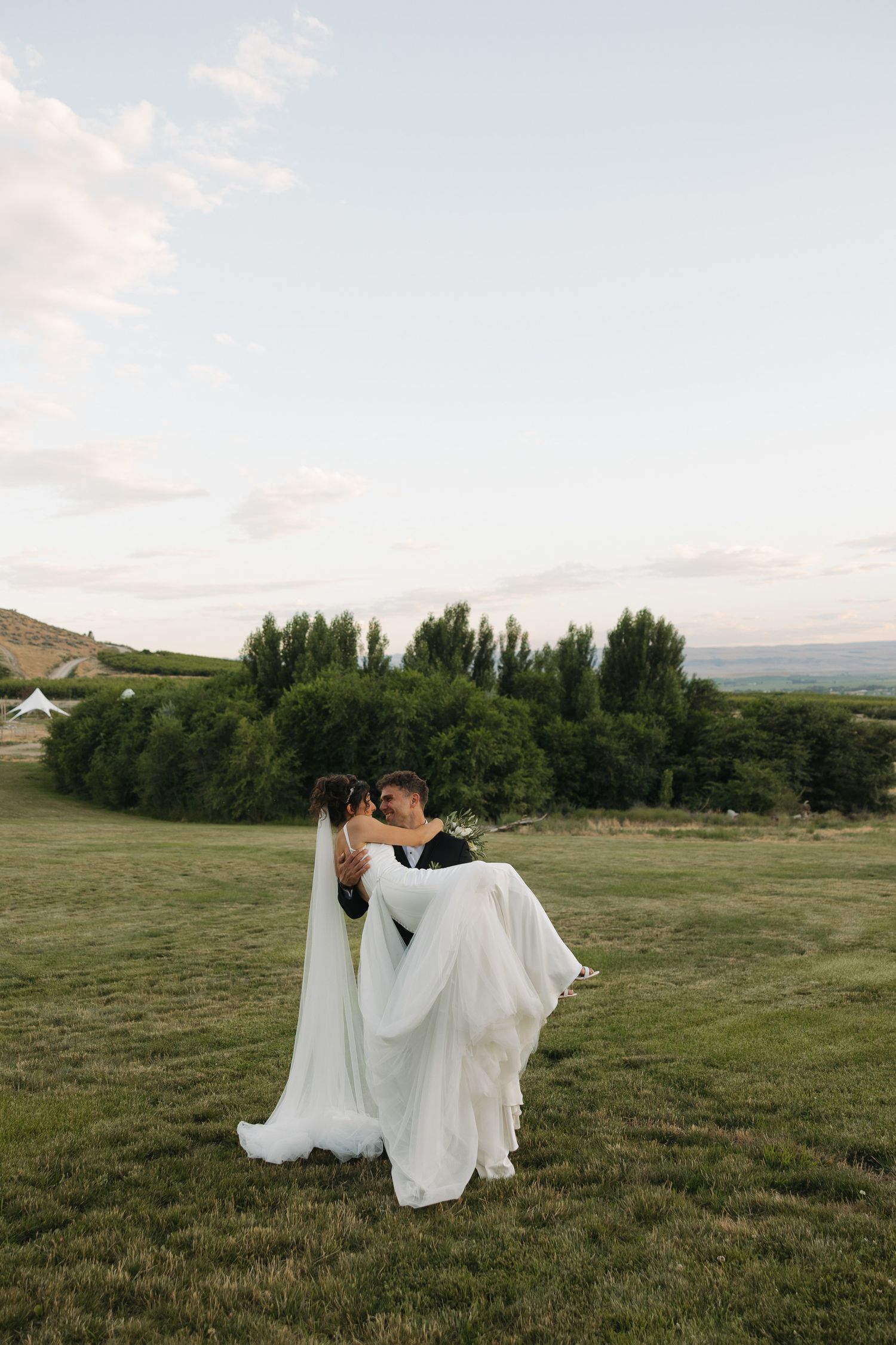 Groom lifts bride in white wedding gown for a kiss in a scenic green field.