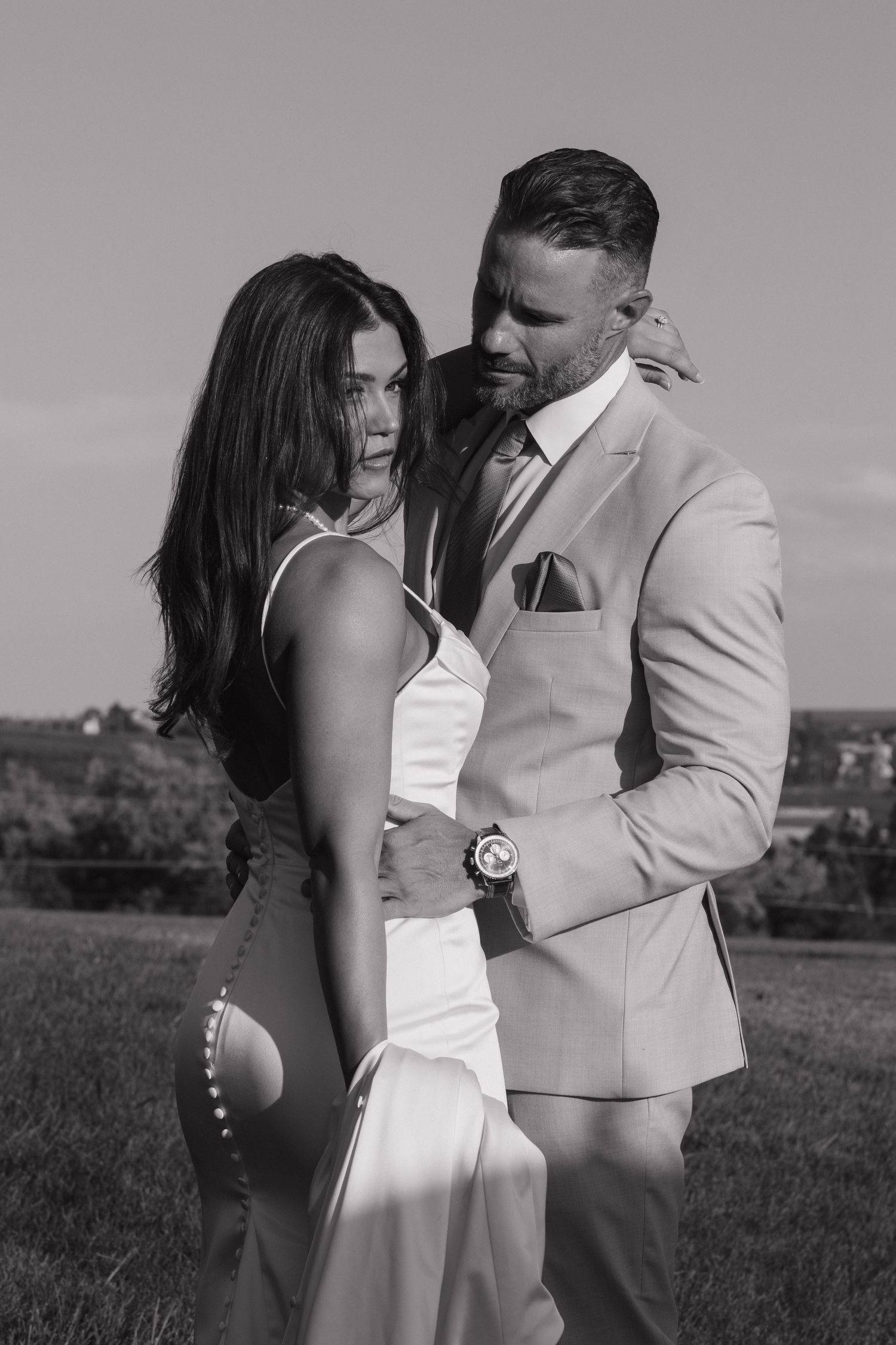 Black and white photo of elegant couple embracing outdoors, woman in white gown and man in beige suit.