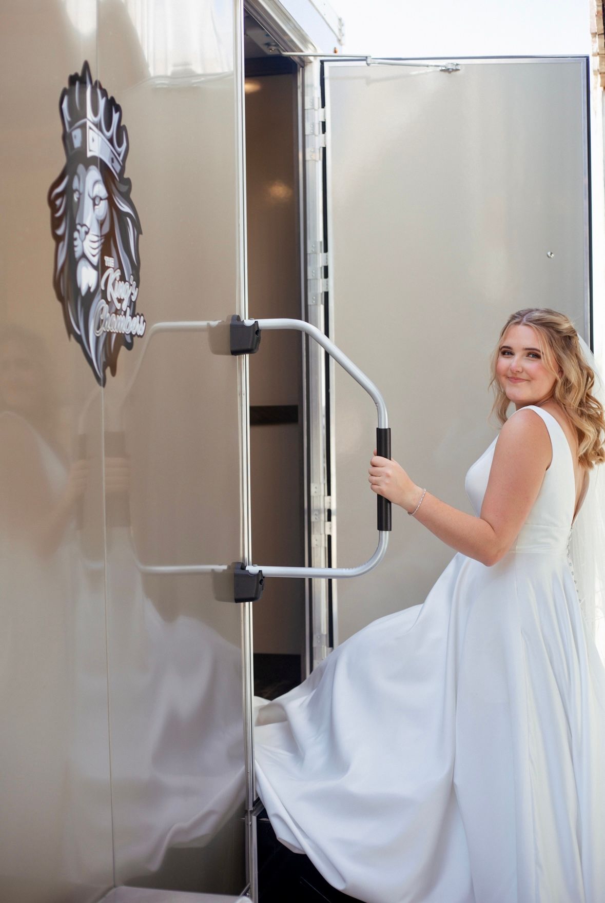 Bride in white gown entering a luxury restroom trailer with lion logo at an outdoor wedding event.
