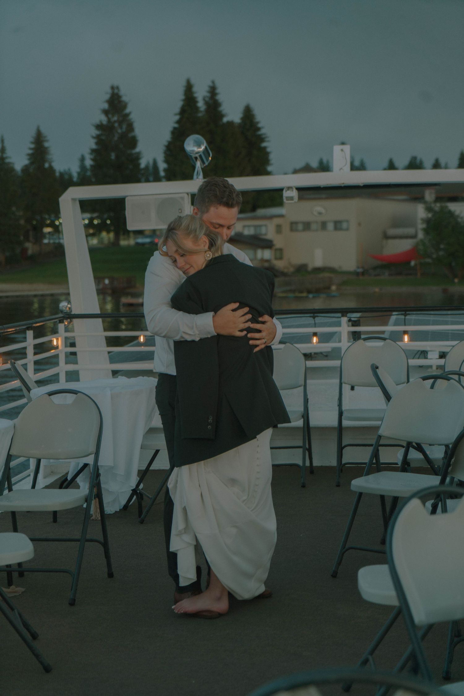 Couple sharing tender embrace while slow dancing on boat deck at dusk with candles and empty chairs nearby.
