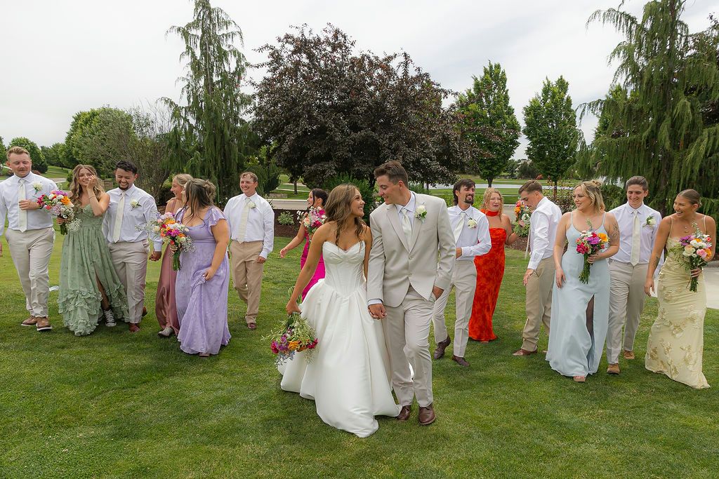 Wedding party walking on green lawn, bride in white gown with groom, bridesmaids in purple and blue dresses, groomsmen in tan suits.