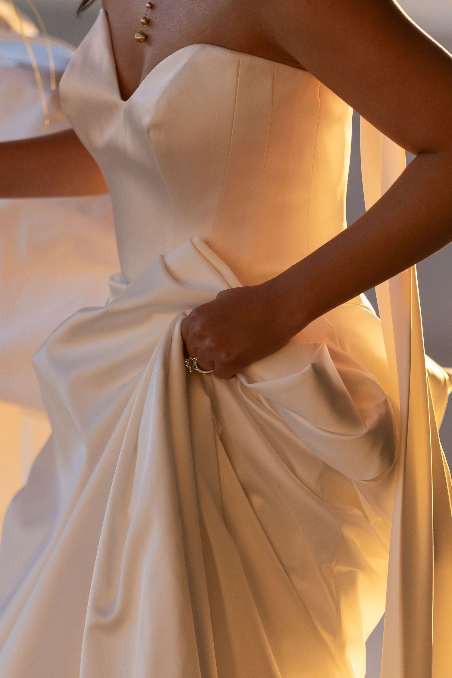 Close-up of a bride in a strapless satin wedding gown, holding the draped skirt in golden sunlight.