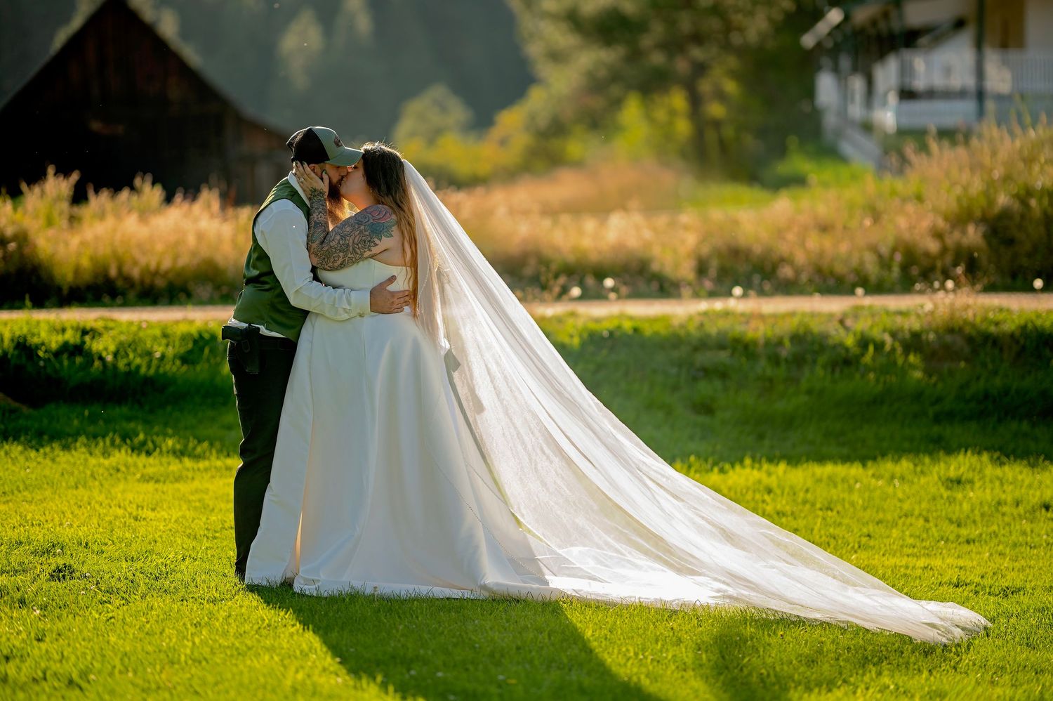 Bride in a flowing white gown with long veil kissing groom in a sunlit green meadow.